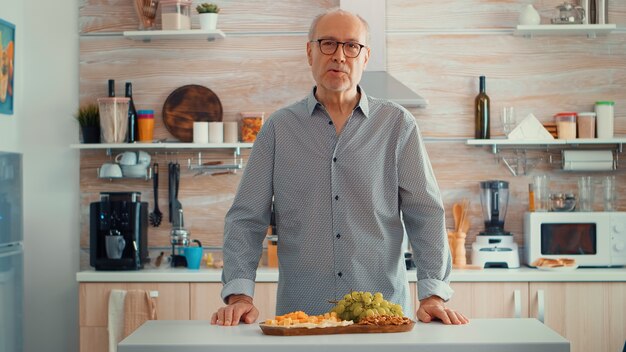 Hombre mayor que tiene una videollamada en la cocina mientras su familia prepara la cena en segundo plano. Punto de vista en línea Internet conferencia moderna, chat, comunicación, conversación de chat llamada a través de cámara web