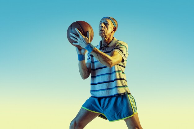 Hombre mayor con ropa deportiva jugando baloncesto sobre fondo degradado, luz de neón. Modelo masculino caucásico en gran forma se mantiene activo. Concepto de deporte, actividad, movimiento, bienestar, confianza.