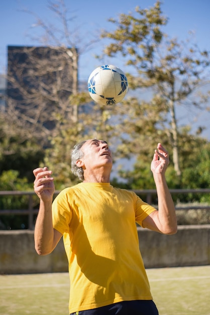 Hombre mayor satisfecho con pelota de fútbol. Hombre caucásico con cabello gris corto en ropa deportiva lanzando pelota en el campo deportivo solo. Fútbol, deporte, concepto de ocio.