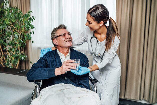 Un hombre mayor en silla de ruedas le sonríe a la asistente de enfermería, ella le entrega un vaso de agua.