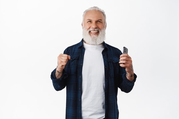 El hombre mayor sonriente dice que sí, levanta el puño, aprieta los puños en señal de triunfo y sostiene el teléfono inteligente, ríe mientras gana y celebra el éxito, logra el objetivo en línea en la aplicación, pared blanca