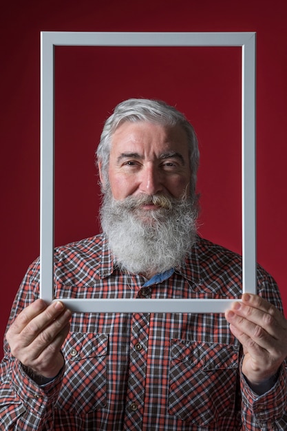 Hombre mayor sonriente que lleva a cabo el marco blanco de la frontera contra fondo coloreado