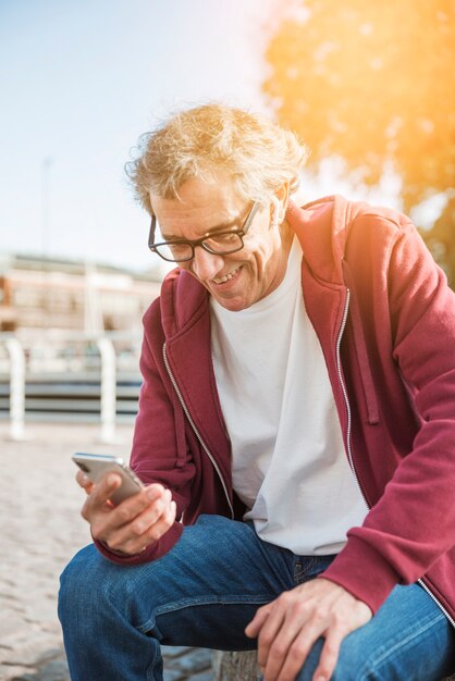 Hombre mayor sonriente que se sienta en el parque que mira smartphone