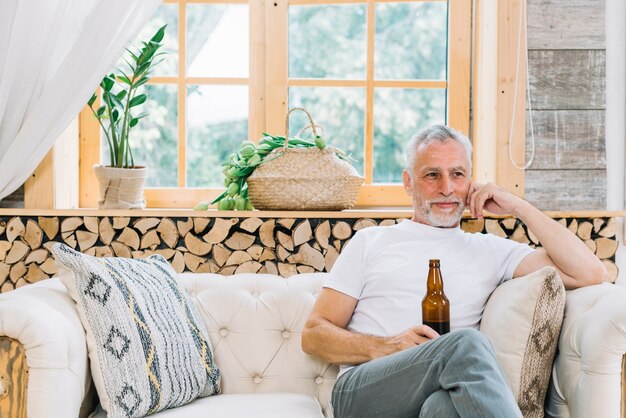 Hombre mayor sonriente que se sienta en el sofá que sostiene la botella de cerveza