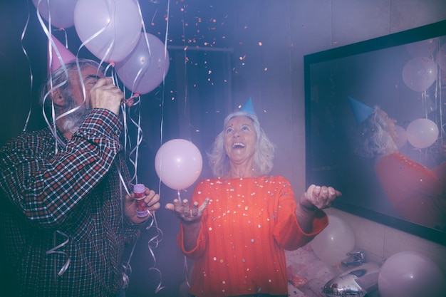 Hombre mayor sonriente que sopla varita de burbuja y su esposa lanzando confeti en la fiesta de cumpleaños