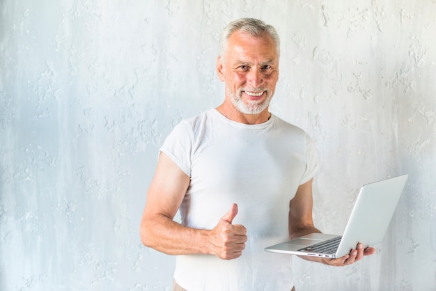 Foto gratuita hombre mayor sonriente que sostiene la computadora portátil que muestra el pulgar encima de muestra