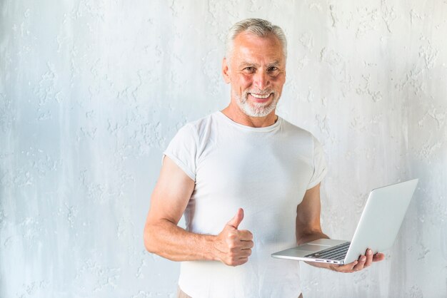 Hombre mayor sonriente que sostiene la computadora portátil que muestra el pulgar encima de muestra