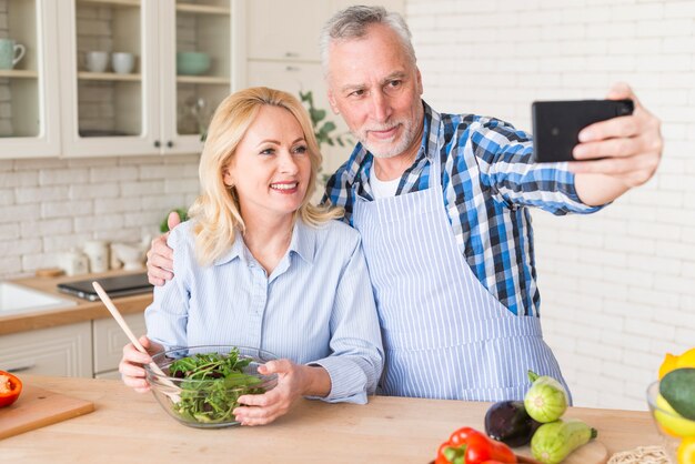 Hombre mayor con su esposa con el tazón de ensalada verde que toma el selfie en el teléfono móvil en la cocina