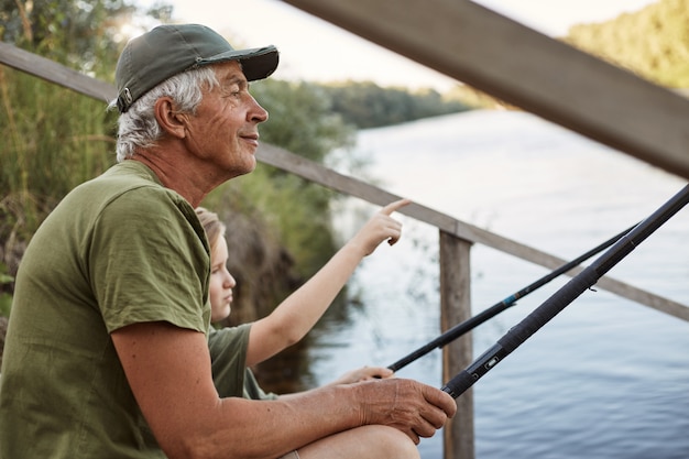 Hombre mayor con su nieto sentado en pontón de madera con cañas de pescar en las manos, disfrutando de la hermosa naturaleza, niño señalando algo con el dedo.