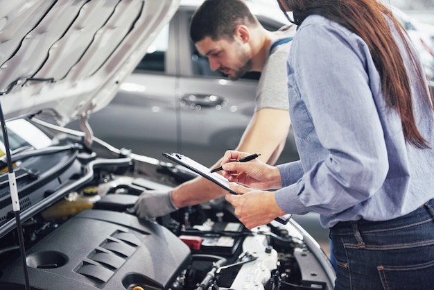 Un hombre mecánico y una mujer cliente miran el capó del automóvil y discuten las reparaciones