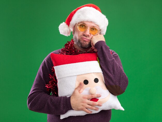 Hombre de mediana edad aburrido con gorro de Papá Noel y guirnalda de oropel alrededor del cuello con gafas sosteniendo la almohada de santa claus manteniendo la mano en la cara aislada en la pared verde