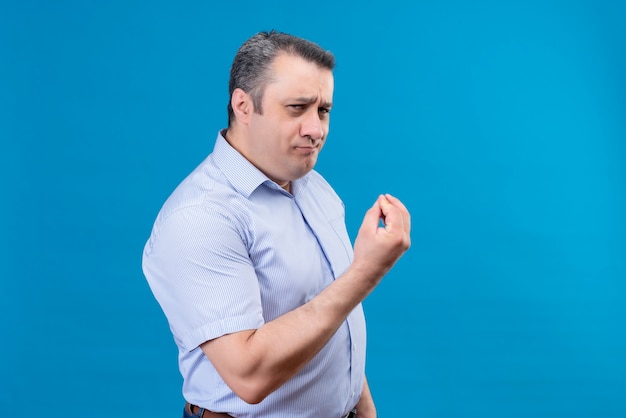 Hombre de mediana edad alegre en camisa azul a rayas verticales mostrando delicioso gesto con la mano sobre un fondo azul.