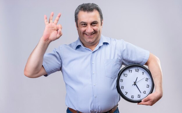 Hombre de mediana edad alegre en camisa azul a rayas verticales sosteniendo un gran reloj haciendo bien firmar con los dedos