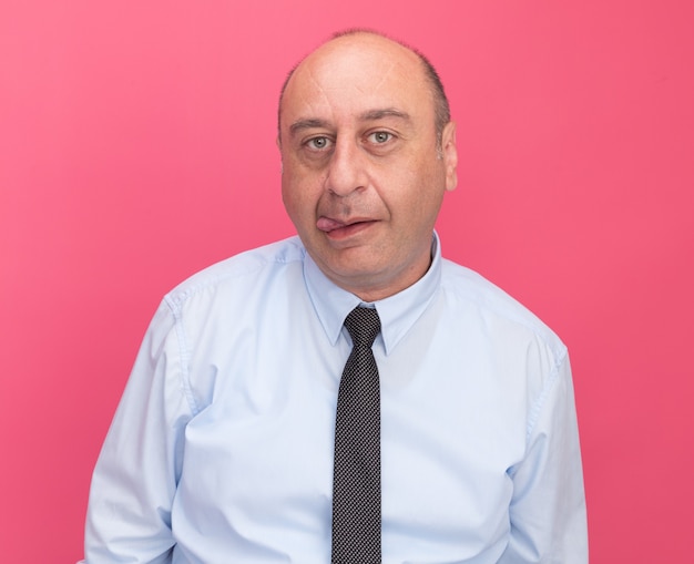 Hombre de mediana edad alegre con camiseta blanca con corbata mostrando la lengua aislada en la pared rosa