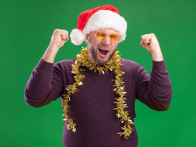 Hombre de mediana edad alegre con gorro de Papá Noel y guirnalda de oropel alrededor del cuello con gafas mirando a la cámara guiñando un ojo haciendo sí gesto aislado sobre fondo verde