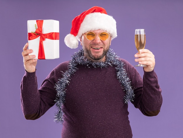 Hombre de mediana edad alegre con gorro de Papá Noel y guirnalda de oropel alrededor del cuello con gafas con paquete de regalo y copa de champán riendo aislado en la pared púrpura