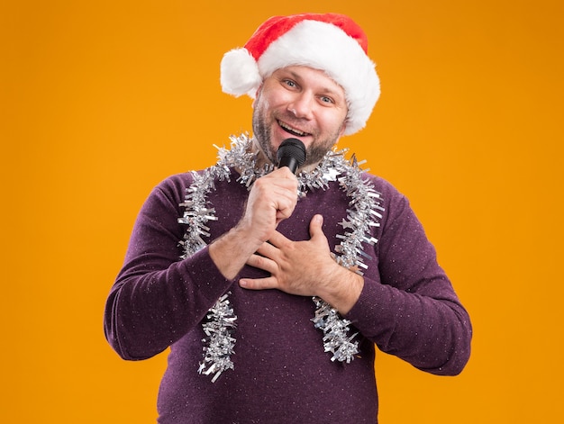 Hombre de mediana edad alegre con gorro de Papá Noel y guirnalda de oropel alrededor del cuello hablando por el micrófono mirando a la cámara manteniendo la mano en el pecho aislado sobre fondo naranja