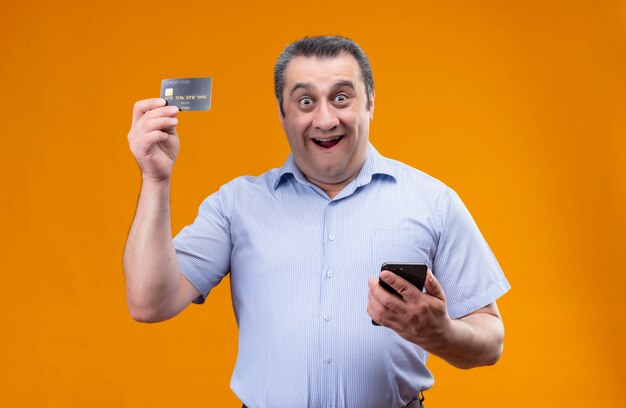 Hombre de mediana edad alegre y positivo con camisa azul a rayas sosteniendo el teléfono móvil y mostrando la tarjeta de crédito mientras está de pie