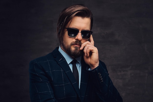 Un hombre de mediana edad, apuesto y pensativo, con barba y peinado, vestido con un elegante traje formal y gafas de sol sobre un fondo oscuro texturizado en el estudio.