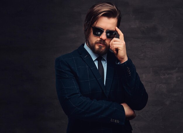 Un hombre de mediana edad, apuesto y pensativo, con barba y peinado, vestido con un elegante traje formal y gafas de sol sobre un fondo oscuro texturizado en el estudio.