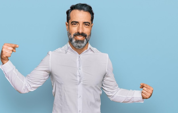 Hombre de mediana edad con barba y camisa blanca casual que se ve confiado con una sonrisa en la cara, apuntándose con los dedos orgulloso y feliz.