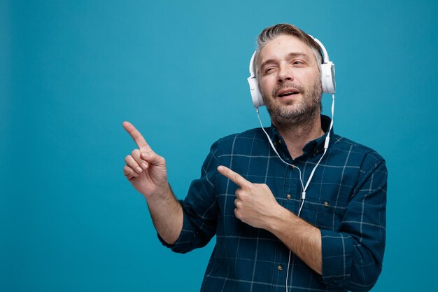 Hombre de mediana edad con cabello gris en camisa de color oscuro con auriculares mirando a un lado feliz y positivo señalando con los dedos índices hacia el lado de pie sobre fondo azul