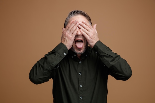 Hombre de mediana edad con cabello gris en camisa de color oscuro feliz y emocionado cubriendo los ojos con las manos sacando la lengua de pie sobre fondo marrón