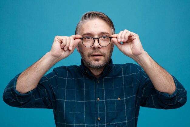 Hombre de mediana edad con cabello gris en camisa de color oscuro con gafas mirando a la cámara con expresión segura de pie sobre fondo azul.