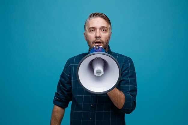 Hombre de mediana edad con cabello gris en camisa de color oscuro hablando en megáfono sorprendido de pie sobre fondo azul
