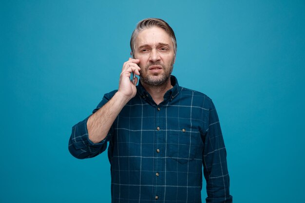 Hombre de mediana edad con cabello gris en camisa de color oscuro hablando por teléfono móvil con aspecto disgustado haciendo la boca torcida de pie sobre fondo azul.