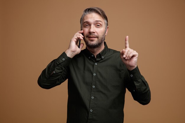 Hombre de mediana edad con cabello gris en camisa de color oscuro hablando por teléfono móvil sonriendo confiado señalando con el dedo índice de pie sobre fondo marrón