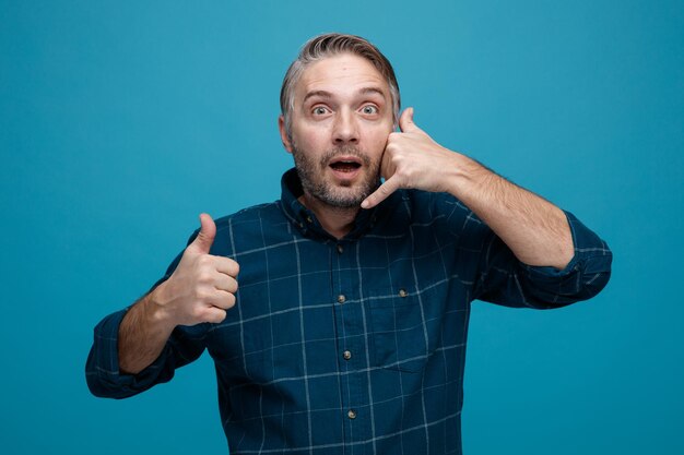 hombre de mediana edad con cabello gris en camisa de color oscuro haciendo un gesto de llamarme mostrando el pulgar hacia arriba feliz y positivo sonriendo de pie sobre fondo azul