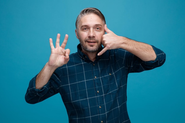 Hombre de mediana edad con cabello gris en camisa de color oscuro haciendo un gesto de llamarme mostrando un signo de ok feliz y positivo sonriendo de pie sobre fondo azul