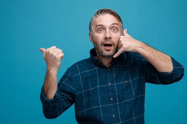 Hombre de mediana edad con cabello gris en camisa de color oscuro mirando a la cámara asombrado y sorprendido mostrando el gesto de llamarme apuntando con el pulgar hacia el lado de pie sobre fondo azul