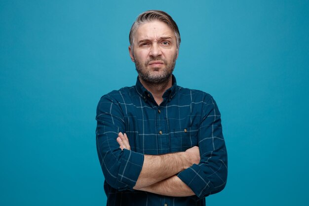 Hombre de mediana edad con cabello gris en camisa de color oscuro mirando a la cámara disgustado haciendo que la boca irónica cruce las manos en el pecho ofendido de pie sobre fondo azul