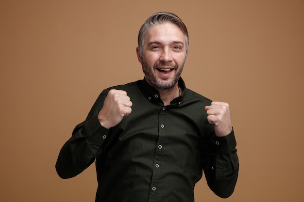 Hombre de mediana edad con cabello gris en camisa de color oscuro mirando a la cámara emocionado y feliz apretando los puños regocijándose de su éxito de pie sobre fondo marrón