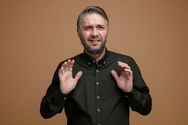 Hombre de mediana edad con cabello gris en camisa de color oscuro mirando a la cámara con expresión repugnante haciendo gesto de defensa con las manos de pie sobre fondo marrón