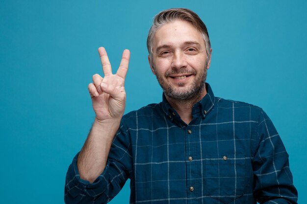 hombre de mediana edad con cabello gris en camisa de color oscuro mirando a la cámara feliz y alegre mostrando vsign sonriendo de pie sobre fondo azul