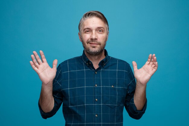 Hombre de mediana edad con cabello gris en camisa de color oscuro mirando a la cámara feliz y positivo levantando los brazos de pie sobre fondo azul.