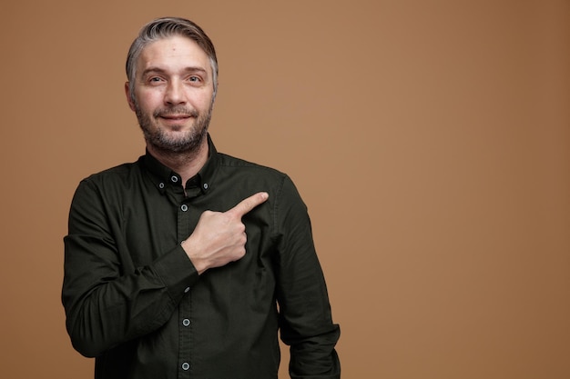 Hombre de mediana edad con cabello gris en camisa de color oscuro mirando a la cámara feliz y positivo señalando con el dedo índice hacia el lado de pie sobre fondo marrón