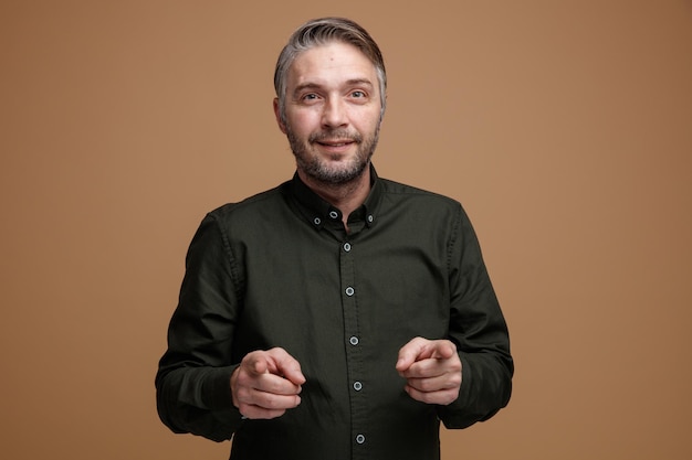 Foto gratuita hombre de mediana edad con cabello gris en camisa de color oscuro mirando a la cámara feliz y positivo señalando con los dedos índices a la cámara de pie sobre fondo marrón