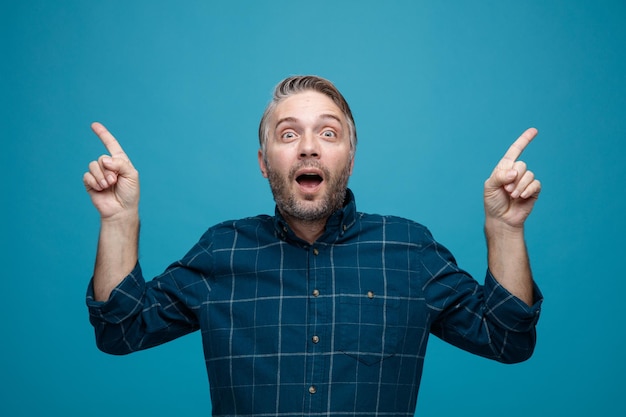 Hombre de mediana edad con cabello gris en camisa de color oscuro mirando a la cámara feliz y sorprendido señalando con los dedos índices de pie sobre fondo azul