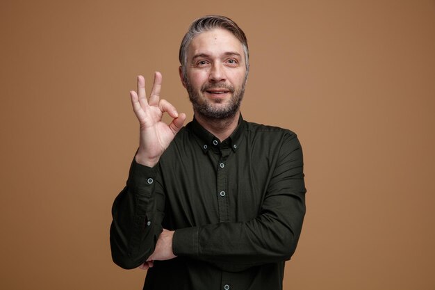 Hombre de mediana edad con cabello gris en camisa de color oscuro mirando a la cámara sonriendo feliz y positivamente mostrando el signo de ok de pie sobre fondo marrón