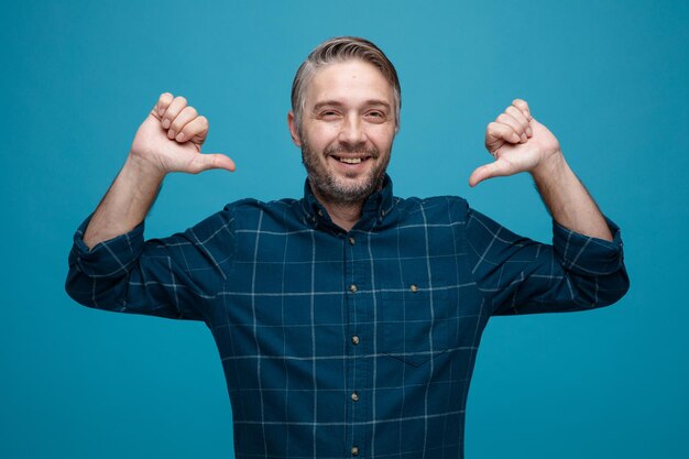 Hombre de mediana edad con cabello gris en camisa de color oscuro mirando a la cámara sonriendo feliz y positivo apuntándose con los pulgares a sí mismo orgulloso y seguro de pie sobre fondo azul