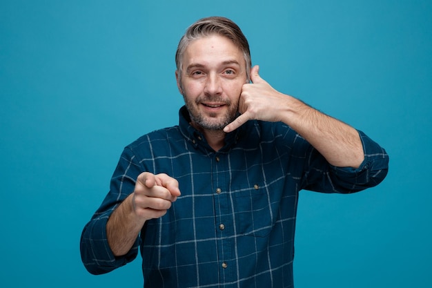 Foto gratuita hombre de mediana edad con cabello gris en camisa de color oscuro mirando a la cámara sonriendo haciendo un gesto de llamarme señalando con el dedo índice a la cámara de pie sobre fondo azul