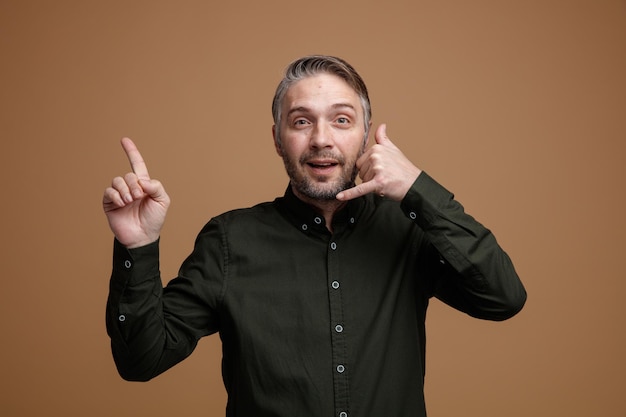 Hombre de mediana edad con cabello gris en camisa de color oscuro mirando a la cámara sonriendo mostrando el gesto de llamarme apuntando con el dedo índice hacia el lado de pie sobre fondo marrón