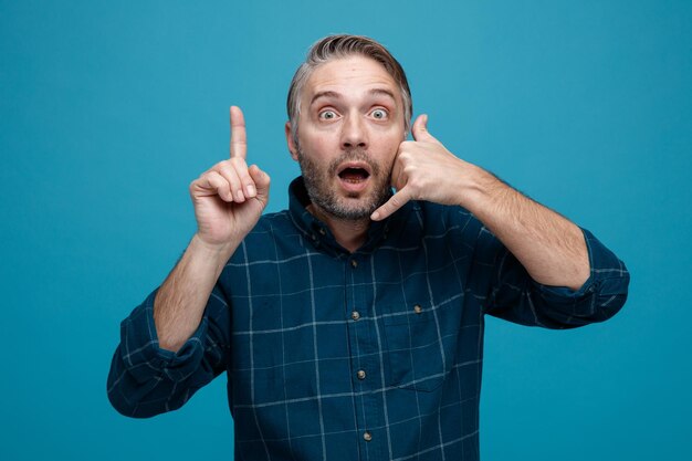 Hombre de mediana edad con cabello gris en camisa de color oscuro mirando a la cámara sorprendido haciendo un gesto de llamarme con la mano apuntando con el dedo índice de pie sobre fondo azul