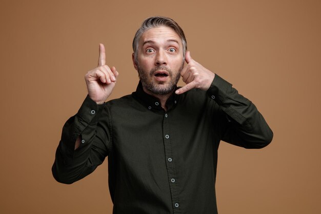 Hombre de mediana edad con cabello gris en camisa de color oscuro mirando a la cámara sorprendido haciendo un gesto de llamarme con la mano apuntando con el dedo índice de pie sobre fondo marrón