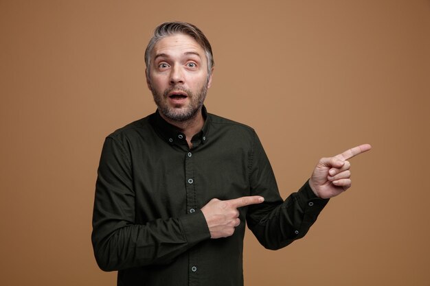 Hombre de mediana edad con cabello gris en camisa de color oscuro mirando a la cámara sorprendido señalando con los dedos índices hacia un lado de pie sobre fondo marrón