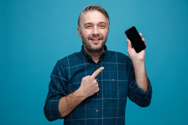 Hombre de mediana edad con cabello gris en camisa de color oscuro que muestra el teléfono inteligente apuntando con el dedo índice mirando a la cámara sonriendo feliz y positivo de pie sobre fondo azul